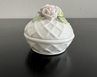 Puede incluir: Una caja de baratijas de cerámica blanca con un patrón de celosía y una tapa en forma de rosa. La rosa tiene pétalos rosas y hojas verdes. La caja es redonda.