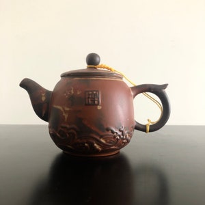 May include: A brown ceramic teapot with a dark brown handle and a dark brown lid. The teapot has a wave design and a small, dark brown knob on the lid. A yellow string is attached to the knob.