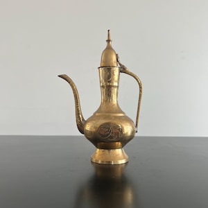 Puede incluir: Una tetera de latón vintage con un diseño ornamentado. La tetera presenta un pico largo y curvo, un asa y una tapa con una parte superior puntiaguda. La superficie está cubierta de patrones grabados, lo que le da una apariencia detallada y antigua.