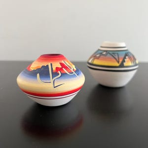 May include: Two small, hand-painted ceramic vases. One vase features a sunset design with a cactus silhouette, and the other has a geometric landscape. Both vases have a white base and are adorned with vibrant colours, showcasing a Southwestern theme.