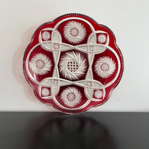 May include: A decorative glass plate in a deep ruby red, with a scalloped edge. The plate features detailed white starburst and geometric patterns. A silver border trims the edge. This elegant plate would make a lovely addition to any home, measuring approximately 20 cm in diameter.