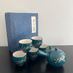 Op de afbeelding: Een turquoise keramische theeset met een theepot en vijf bijpassende kopjes. Elk stuk heeft een bloemmotief in rood, geel en groen. Een blauwe doos met Japanse tekst staat op de achtergrond.