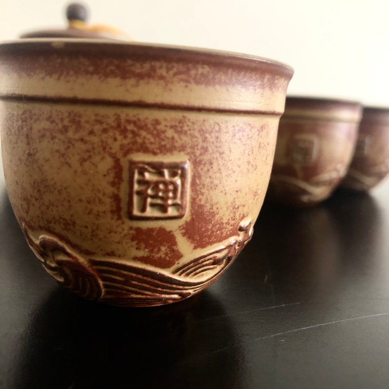 May include: A set of four brown ceramic teacups with a wave design and a square character in the center of each cup. The character on the front cup is "禪".