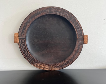 May include: A round, dark brown wooden tray with carved geometric patterns around the rim. The tray has two small, rectangular handles and a smooth, dark interior. The tray is displayed on a black surface against a white wall.