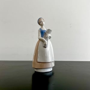 May include: A porcelain figurine of a woman wearing a blue and white dress and a blue hat, holding a bouquet of flowers.