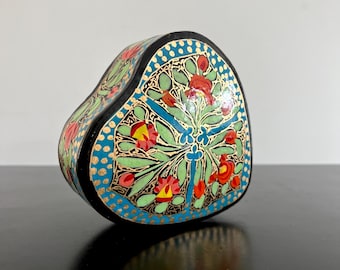 May include: Heart-shaped decorative box with a black base and lid. The box is adorned with a floral design featuring red, yellow, and green flowers, accented by blue and gold details. The box is sitting on a black surface.