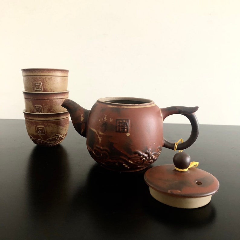 May include: A brown ceramic teapot with a lid and three matching cups. The teapot has a decorative design of waves and a square symbol. The cups are stacked on top of each other.