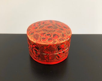 Pode incluir: Uma pequena caixa redonda vermelha com um desenho floral. A caixa é pintada com detalhes pretos e dourados.