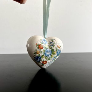 May include: Cream-coloured heart-shaped ornament with a floral design in blue, orange, and yellow. A pale green ribbon is attached for hanging. The ornament is likely ceramic or porcelain.