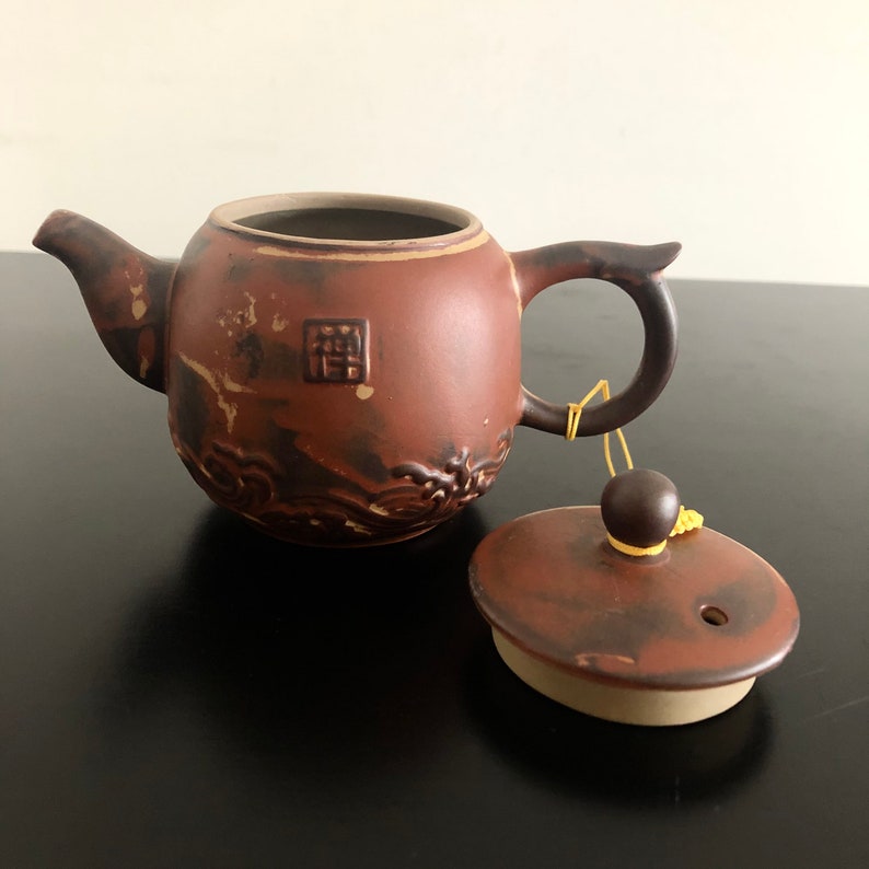 May include: A brown ceramic teapot with a lid and a carved design. The teapot has a small handle and a spout. The lid has a small knob on top. The teapot is sitting on a black surface.
