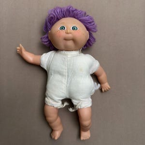 May include: A vintage Cabbage Patch Kid doll with purple yarn hair, green eyes, and a white onesie. The doll has a rosy complexion and a sweet expression.
