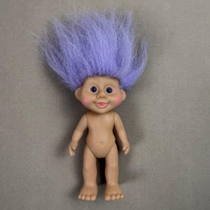 May include: A vintage troll doll with a light skin tone, blue eyes, and a smiling expression. The doll has a tuft of purple hair and rosy cheeks. The doll is standing upright with arms at its sides.