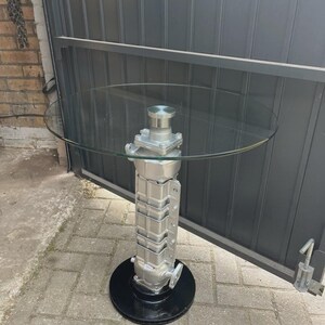 May include: A circular glass-topped table featuring a silver-coloured automotive engine component as a central pedestal base, which is mounted onto a solid black circular metal disc that serves as the floor support for the entire structure.