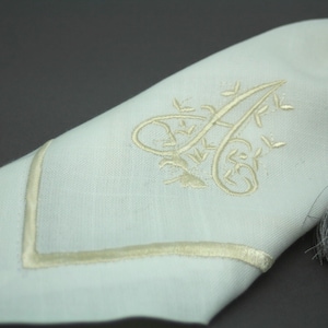 May include: A white linen napkin with a gold embroidered letter "A" and a gold border. The napkin has a grey tassel.