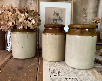 May include: Three vintage brown and white ceramic jars with a brown rim and a white body. The jars are sitting on a wooden table with a newspaper underneath.