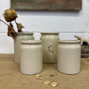 May include: Four small, off-white ceramic jars with a speckled finish. The jars are cylindrical with a slightly flared rim. One jar has faded floral accents. A dried flower and leaves are in one jar. The jars are on a burlap surface.