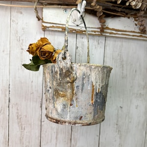May include: A weathered metal bucket with a worn, white paint finish. The bucket has a handle and is decorated with dried flowers.