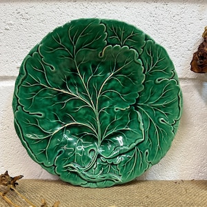 May include: A green ceramic plate, shaped like a large leaf, with intricate veining and a glossy finish. The plate's design is a rich shade of green, bringing a touch of nature-inspired elegance to any table setting. The plate is approximately 30 cm in diameter.