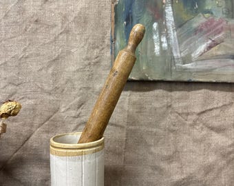 May include: A cylindrical white ceramic jar with vertical ribbed texture and a tan rim, containing a long, slender, weathered wooden rolling pin that extends diagonally upward from the opening of the container.