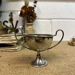May include: A small, silver cup with two curved handles. The cup has a rounded bowl shape and sits on a pedestal base. The surface of the cup reflects light, and the overall design is vintage.