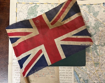 May include: A fabric Union Jack flag with the text "BRITISH MADE" printed in the corner, attached to a wooden pole. The flag rests on top of a vintage map, a dark green rectangular object, and a piece of paper with handwritten cursive text.