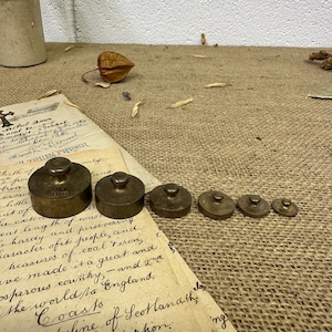 May include: A set of antique brass weights of different sizes, displayed on a textured surface with old paper and a dried seed pod. The weights are circular with a small knob on top.