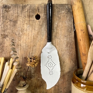 May include: White enamel slotted spoon with a black handle. The spoon is leaf-shaped and has small holes. The spoon is on a wooden chopping board.