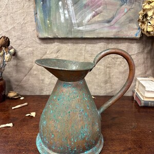 May include: A copper pitcher with a wide, flared spout and a curved handle. The metal surface displays a weathered patina with visible oxidation, featuring a mottled texture of brown copper tones mixed with patches of bright teal and green.