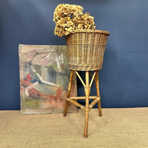 May include: A vintage wicker plant stand featuring a woven basket and a tripod base. The stand is made of light brown wood and holds dried flowers. An abstract painting leans against a dark blue wall.