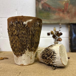 May include: Two handcrafted drums with a natural, rustic aesthetic. The drums have brown and white animal hide tops and bases, with woven, light brown sides. One drum is upright, the other is on its side.