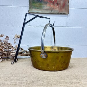 May include: A vintage brass pot with a silver handle. The pot has a worn, aged look and is hanging from a black metal hook.