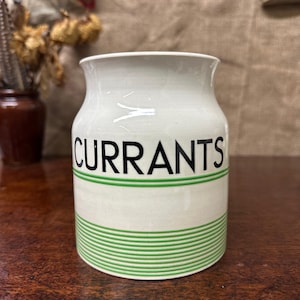 May include: A white ceramic jar with the word "CURRANTS" in black lettering. The jar has a green striped band around the base. The jar is likely used for storing food items.