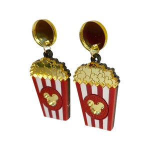 Puede incluir: Un par de pendientes novedosos con dise&ntilde;os de cubos de palomitas de ma&iacute;z. Los pendientes tienen cubos a rayas rojas y blancas con un c&iacute;rculo rojo y una cabeza dorada de Mickey Mouse. Las palomitas son doradas y los postes de los pendientes son dorados.