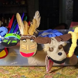 May include: Five sock puppets in a clear plastic bin. The puppets are made from socks and have different faces and features. One puppet has a brown face with a red mouth and feathers on its head. Another puppet has a black face with a yellow beak and large eyes. A third puppet has a brown face with a black nose and button eyes. A fourth puppet has a green face with a black nose and green eyes. The fifth puppet has a black face with a red nose and blue and red stripes on its neck.