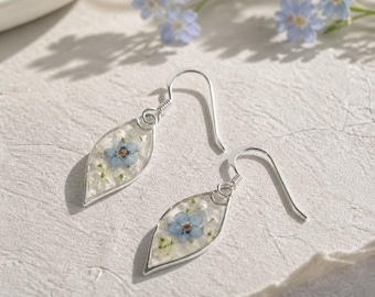 May include: A pair of silver drop earrings featuring a leaf-shaped pendant. Each pendant contains a clear resin inlay with a single pressed light blue forget-me-not flower and small white petals, suspended from a simple silver hook wire.