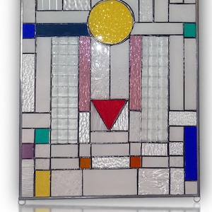 Op de afbeelding: Een rechthoekig glas-in-loodpaneel met een geometrische compositie van heldere, getextureerde glaspanelen afgewisseld met levendige accenten in blauw, geel, rood, groen en paars. Het ontwerp bevat cirkels, driehoeken en rechthoeken.