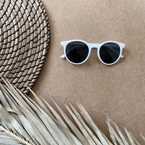 May include: White round sunglasses with black lenses on a brown background with a woven mat and a dried palm leaf.