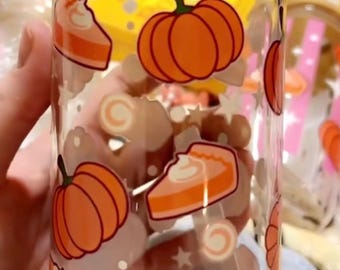 May include: A clear glass jar with a wooden lid, decorated with orange pumpkins, pie slices, and flowers. The jar is perfect for storing small items or as a decorative piece.