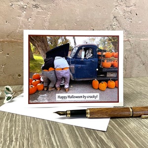 May include: A Halloween greeting card featuring a humorous image of two people with their pants down, working on a truck, with a skeleton driving. The truck bed is filled with pumpkins. The card reads "Happy Halloween by cracky!"