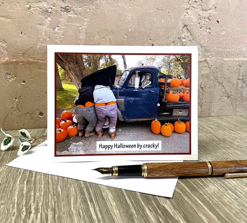 May include: A Halloween greeting card featuring a humorous image of two people with their pants down, working on a truck, with a skeleton driving. The truck bed is filled with pumpkins. The card reads "Happy Halloween by cracky!"