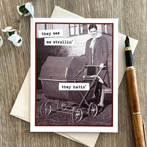 May include: Greeting card with a vintage black and white image of a person pushing a baby carriage. Text on the card reads "they see me strollin' they hatin'". A pen and envelope are next to the card.