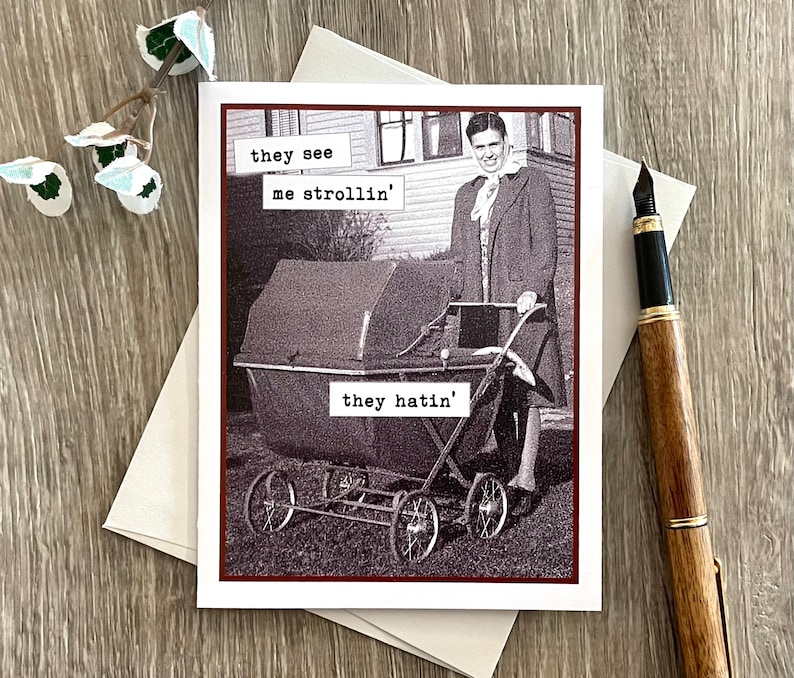 May include: Greeting card with a vintage black and white image of a person pushing a baby carriage. Text on the card reads "they see me strollin' they hatin'". A pen and envelope are next to the card.