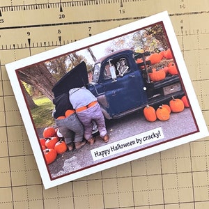 May include: A Halloween-themed greeting card featuring a humorous image. Two people are bent over, working on a vintage blue truck, with a skeleton in the driver's seat. Pumpkins surround the truck. The text "Happy Halloween by cracky!" is displayed.
