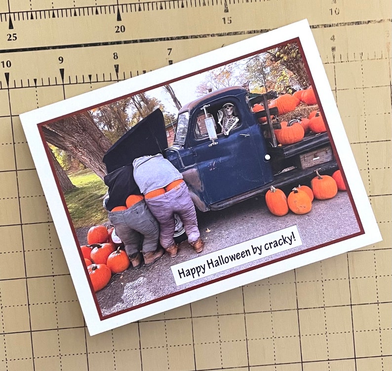 May include: A Halloween-themed greeting card featuring a humorous image. Two people are bent over, working on a vintage blue truck, with a skeleton in the driver's seat. Pumpkins surround the truck. The text "Happy Halloween by cracky!" is displayed.