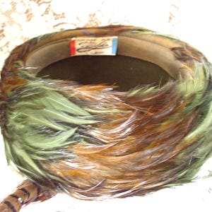May include: A vintage hat featuring a dark interior and a brim decorated with green, brown, and tan feathers. The feathers are arranged to create a full, textured effect. A tag with text is visible on the interior.