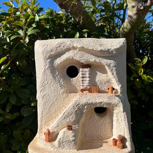 May include: A beige, textured birdhouse with a unique, cave-like design. It features a round entry hole and several small, decorative figurines. The birdhouse is mounted on a wooden post, with green foliage and a blue sky in the background.