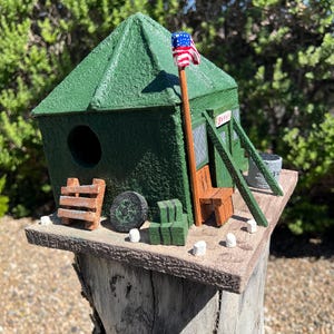 May include: A green birdhouse with a textured roof and walls, featuring a circular entrance hole, a small wooden bench, a miniature American flag on a pole, a ladder, a tire, and stacked blocks on a flat base, with the word SWAMP visible on a sign.