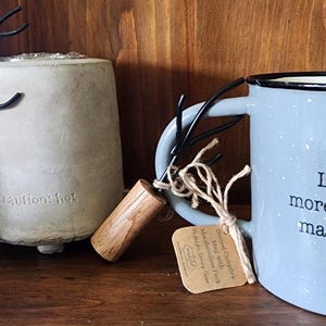 Puede incluir: Una taza de esmalte azul claro moteado con el texto Life needs more chocolate & marshmallows, junto a un tenedor para malvaviscos de metal con mango de madera y una base de hormigón con el texto en relieve caution: hot.