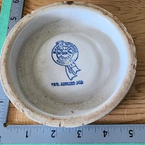 May include: A vintage, off-white ceramic bowl with a blue and white emblem in the center. The emblem reads "Eighty-Five Potter Co. Blue Ribbon Ware" and "Pat. Applied For". The rim of the bowl has a textured, aged appearance.