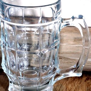 Puede incluir: Una jarra de cerveza de vidrio transparente con asa. La jarra tiene un diseño facetado en el cuerpo y una base robusta. El asa es curva y está unida al lateral. La jarra está vacía y descansa sobre una superficie de madera.